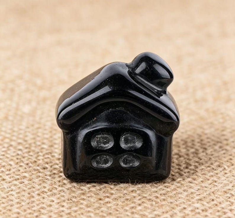 May include: A small, glossy black house figurine with four circular window details. The roof has a curved design, and the overall shape is compact and detailed. The figurine is set against a neutral, textured background.