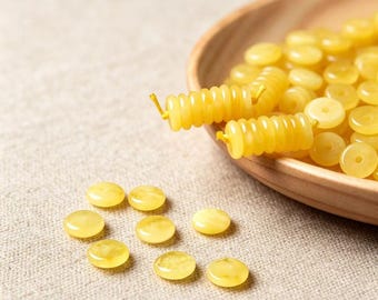May include: A collection of small, round, yellow beads with a smooth, translucent finish. Some beads are loose and scattered, while others are strung together on a thin yellow cord, creating a ribbed, cylindrical shape in a wooden bowl.