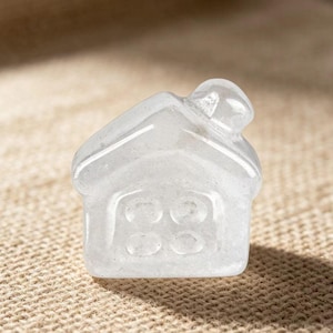 May include: A clear, house-shaped crystal figurine. The miniature house has a pitched roof and a door with three circular windows. The crystal is translucent, allowing light to pass through, and sits on a textured, tan surface.