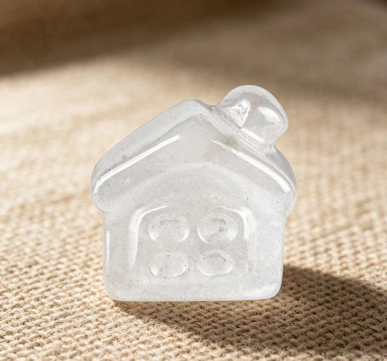 May include: A clear, house-shaped crystal figurine. The miniature house has a pitched roof and a door with three circular windows. The crystal is translucent, allowing light to pass through, and sits on a textured, tan surface.