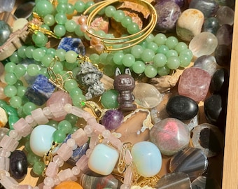 May include: A collection of gemstones and jewelry, including green jade bead necklaces, gold bracelets, and various polished stones in shades of pink, purple, and gray. A small, carved figurine is also present.