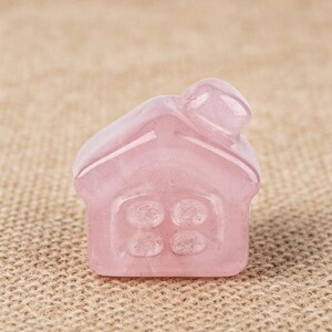 May include: A small, pink rose quartz house figurine. The house has a chimney and four window panes. The gemstone is translucent and has a smooth, polished surface. The figurine is set against a neutral background.