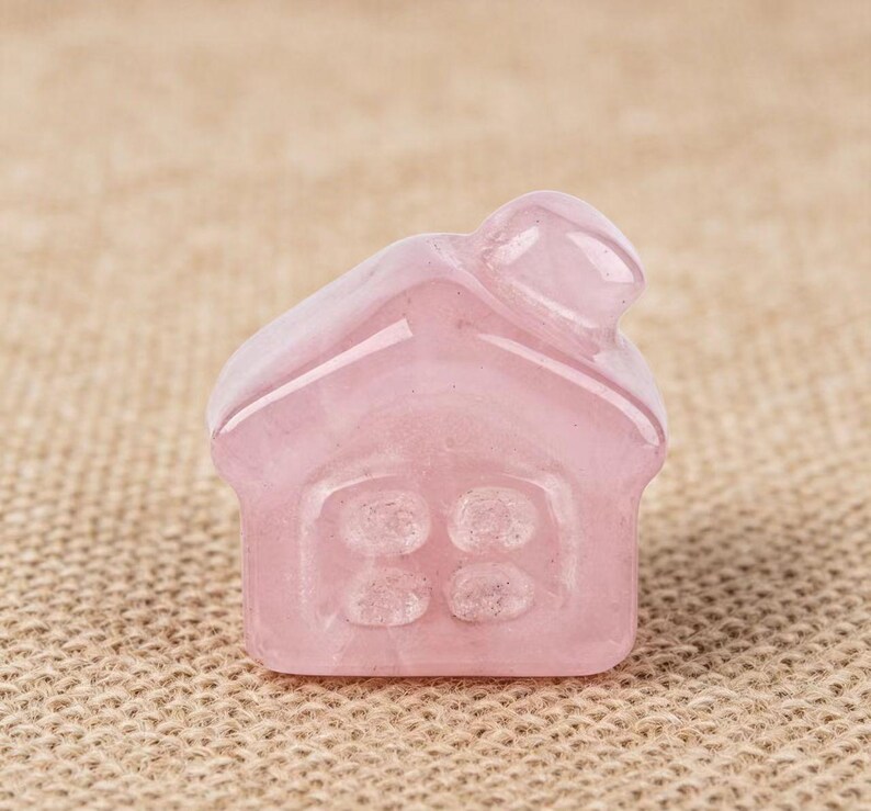 May include: A small, pink rose quartz house figurine. The house has a chimney and four window panes. The gemstone is translucent and has a smooth, polished surface. The figurine is set against a neutral background.