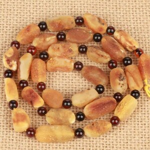May include: A necklace featuring amber-colored beads interspersed with dark brown and red beads. The amber beads vary in shape and size, with a polished, natural appearance. The necklace is strung on a yellow cord.