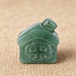 May include: A small, green, gemstone house figurine. The house has a pitched roof and four circular windows on the front. The gemstone has a translucent quality, with visible inclusions. The figurine is set against a neutral, textured background.