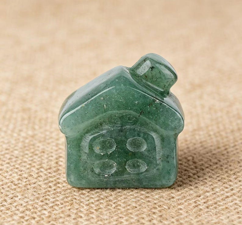 May include: A small, green, gemstone house figurine. The house has a pitched roof and four circular windows on the front. The gemstone has a translucent quality, with visible inclusions. The figurine is set against a neutral, textured background.