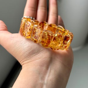 May include: A golden amber bracelet with rectangular, translucent beads. The beads have a warm, honey-like color with reddish-brown inclusions. The bracelet is held in a hand, showcasing its polished, natural appearance.