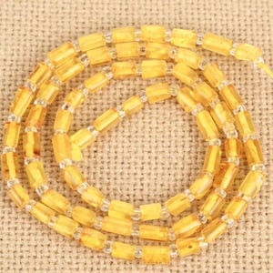 May include: A necklace featuring strands of amber-colored, rectangular beads interspersed with clear, small beads. The beads are strung together, creating a layered effect. The necklace is displayed on a textured, tan surface.