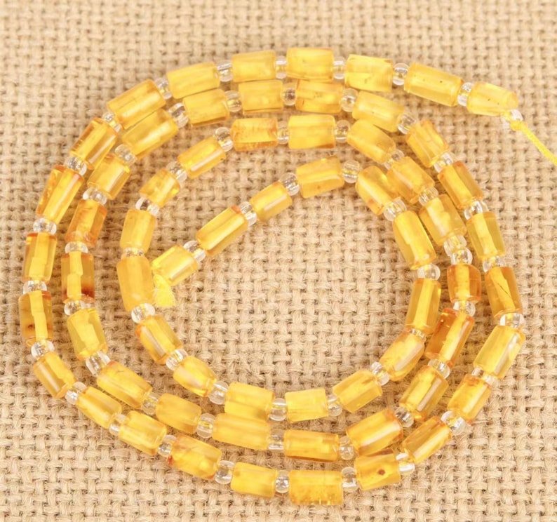 May include: A necklace featuring strands of amber-colored, rectangular beads interspersed with clear, small beads. The beads are strung together, creating a layered effect. The necklace is displayed on a textured, tan surface.