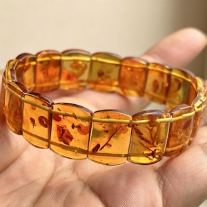 May include: A stretch bracelet composed of rectangular amber beads. The translucent, golden-brown beads contain visible inclusions. The bracelet is strung together with thin, gold-colored wire, creating a flexible design.