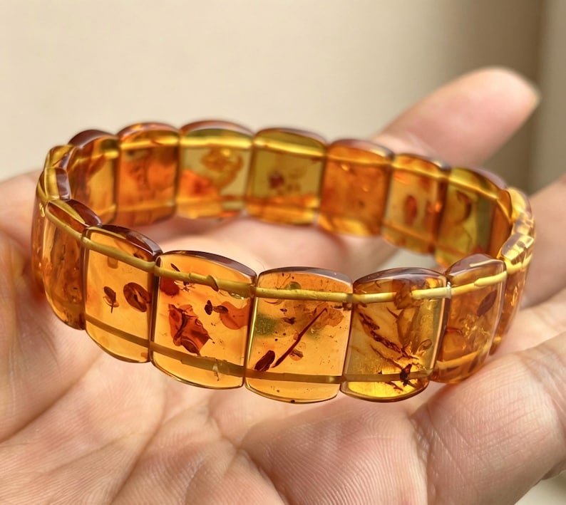 May include: A stretch bracelet composed of rectangular amber beads. The translucent, golden-brown beads contain visible inclusions. The bracelet is strung together with thin, gold-colored wire, creating a flexible design.