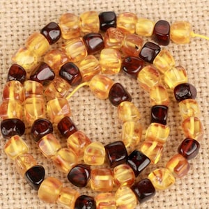 May include: A strand of amber beads in various shades of yellow and brown. The beads are square-shaped and strung together on a yellow string, creating a necklace or bracelet. The amber beads have a polished, glossy appearance.