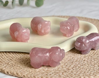 May include: A collection of six pink rose quartz pig figurines. The pig sculptures are displayed on a cream-colored tray and a woven mat. The figurines are carved with detailed features and have a polished, glossy finish.