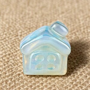 May include: A small, iridescent, house-shaped crystal figurine. The crystal is primarily white with blue and yellow hues. The house has a pitched roof and four circular window details on the front. The figurine is set against a neutral, textured background.