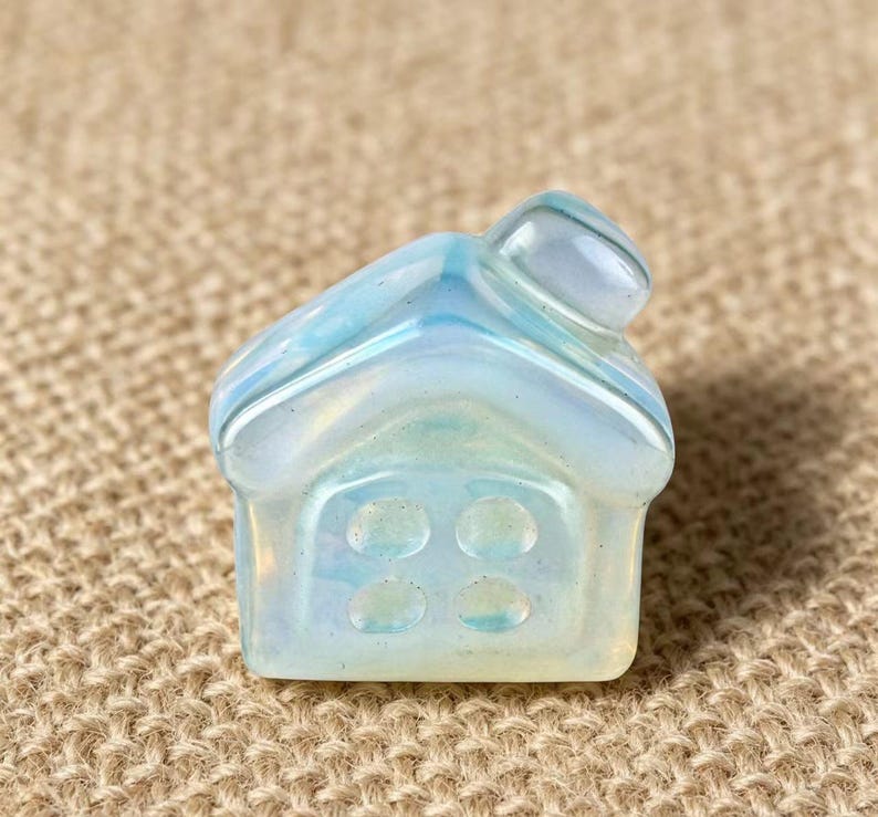 May include: A small, iridescent, house-shaped crystal figurine. The crystal is primarily white with blue and yellow hues. The house has a pitched roof and four circular window details on the front. The figurine is set against a neutral, textured background.