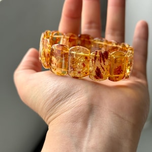 May include: A bracelet made of amber-colored rectangular beads. The beads have a translucent quality with reddish-brown inclusions. The bracelet is displayed on a hand, showcasing its warm tones and unique patterns. A piece of jewelry.