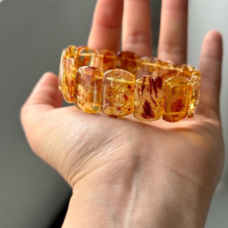 May include: A bracelet made of amber-colored rectangular beads. The beads have a translucent quality with reddish-brown inclusions. The bracelet is displayed on a hand, showcasing its warm tones and unique patterns. A piece of jewelry.
