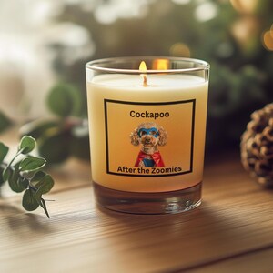 May include: A lit candle in a clear glass jar. The candle has a label with the word "Cockapoo" above an image of a dog in a superhero costume. The text "After the Zoomies" is below the image. The candle sits on a wooden surface.