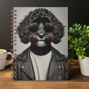 Pode incluir: Um caderno de espiral com a ilustração de um cão preto de pelo encaracolado a usar óculos de sol redondos, uma t-shirt branca e um casaco de cabedal preto. O design está centrado num fundo cinzento claro com uma espiral de cor cobre.