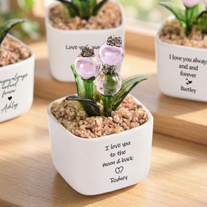 May include: White ceramic pots with glass flower sculptures. Each pot has a different message, such as "I love you to the moon & back" and "I love you always and forever." The glass flowers are pink and green.