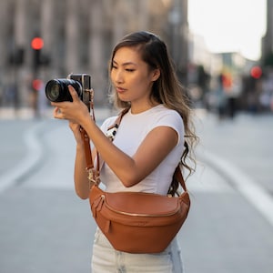 Puede incluir: Un bolso de cintura de cuero marrón con cremallera y correa doradas. La bolsa se lleva cruzada. La persona sostiene una cámara negra con una correa marrón. El fondo es una calle de la ciudad.