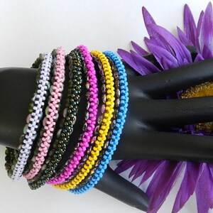 May include: A stack of colorful beaded bracelets in shades of white, pink, green, purple, yellow, and blue. The bracelets are made of small, round beads and are arranged in a circular pattern.