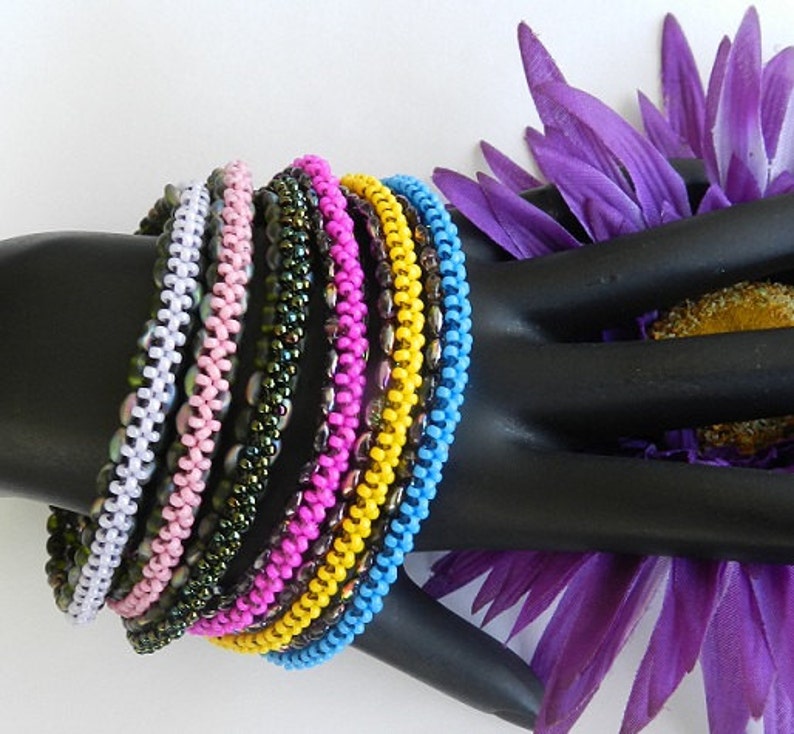 May include: A stack of colorful beaded bracelets in shades of white, pink, green, purple, yellow, and blue. The bracelets are made of small, round beads and are arranged in a circular pattern.