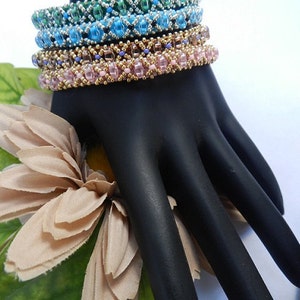 May include: A set of four beaded bracelets in shades of green, blue, pink, and gold. The bracelets are stacked on a black hand model.