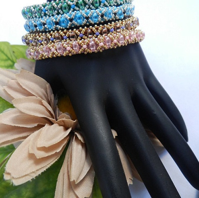 May include: A set of four beaded bracelets in shades of green, blue, pink, and gold. The bracelets are stacked on a black hand model.