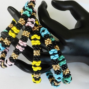 Op de afbeelding: Vier kralenarmbanden in zwart, geel, roze en turquoise met gouden accenten. De armbanden zijn gestapeld op een zwart handmodel.