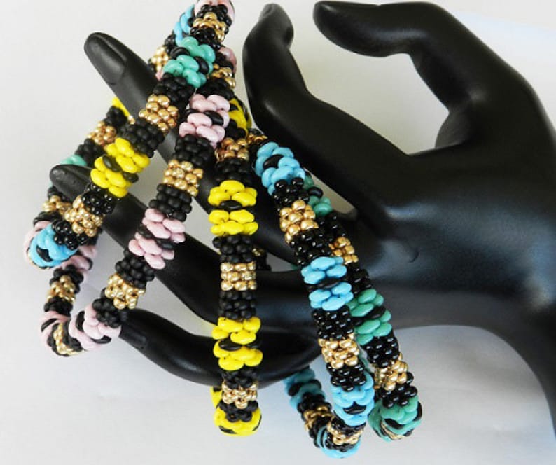 Op de afbeelding: Vier kralenarmbanden in zwart, geel, roze en turquoise met gouden accenten. De armbanden zijn gestapeld op een zwart handmodel.