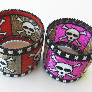 Puede incluir: Dos pulseras de cuentas con un dise&ntilde;o de calavera y huesos cruzados. Una pulsera es roja, dorada y negra, y la otra es rosa, dorada y negra.