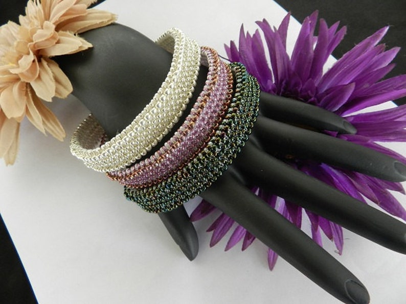 May include: Four beaded bracelets stacked on a black hand model. The bracelets are made of silver, pink, copper, and green beads.