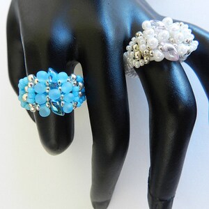May include: Two beaded rings on a black mannequin hand. The ring on the index finger is made of light blue beads, while the ring on the middle finger is made of white and light pink beads.