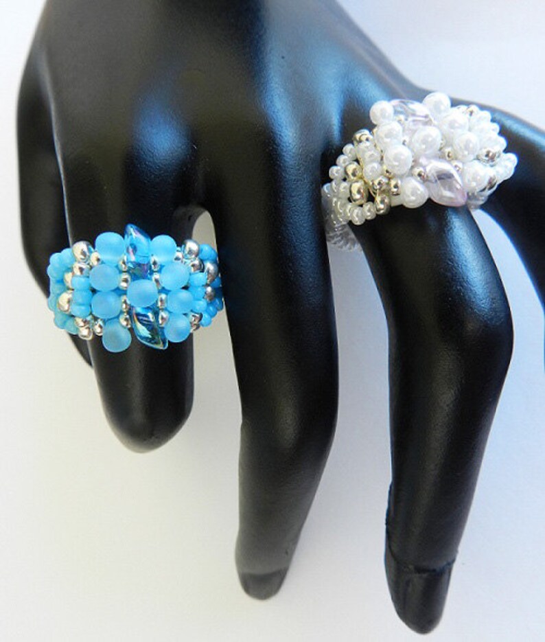 May include: Two beaded rings on a black mannequin hand. The ring on the index finger is made of light blue beads, while the ring on the middle finger is made of white and light pink beads.