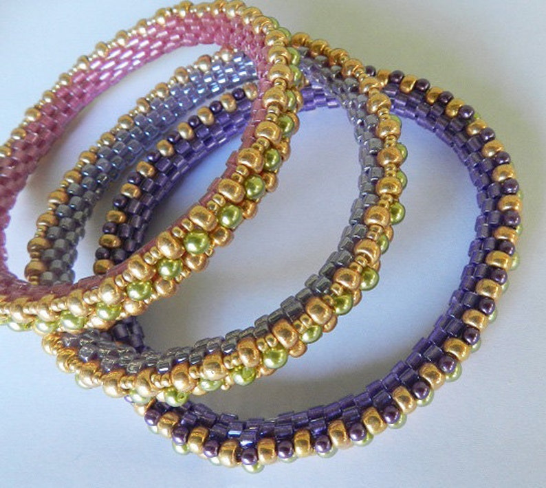 May include: Three beaded bracelets in shades of purple, green, and pink. The bracelets are made with small, round beads and are stacked on top of each other.