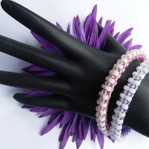 May include: Two beaded bracelets, one pink and white, the other lavender and white, on a black hand model with a purple flower behind it.