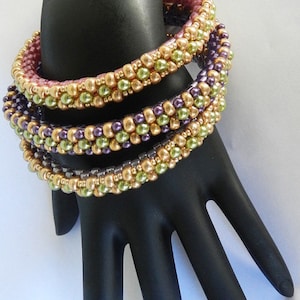 May include: Four beaded bracelets stacked on a black hand model. The bracelets are made with small, round beads in shades of gold, green, and purple.