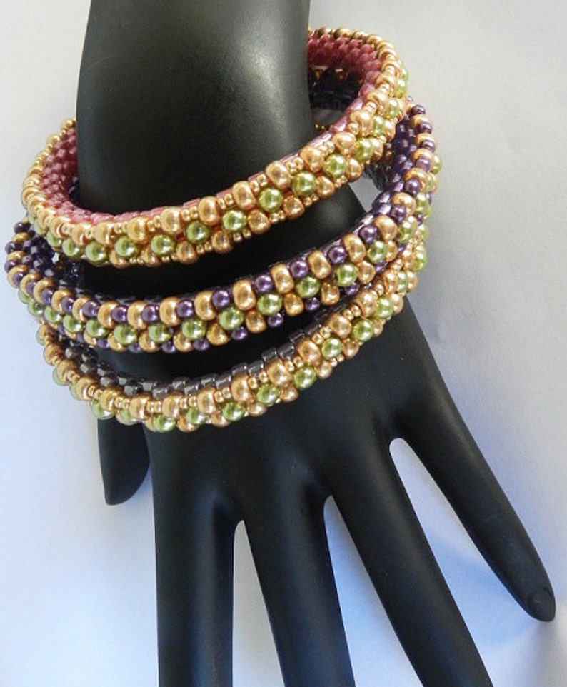 May include: Four beaded bracelets stacked on a black hand model. The bracelets are made with small, round beads in shades of gold, green, and purple.