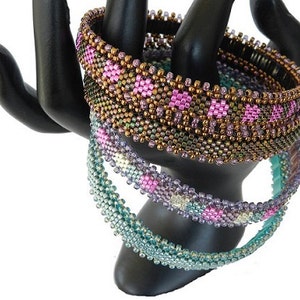 May include: Three beaded bracelets in shades of teal, purple, and brown with pink accents. The bracelets are stacked on a black display stand.