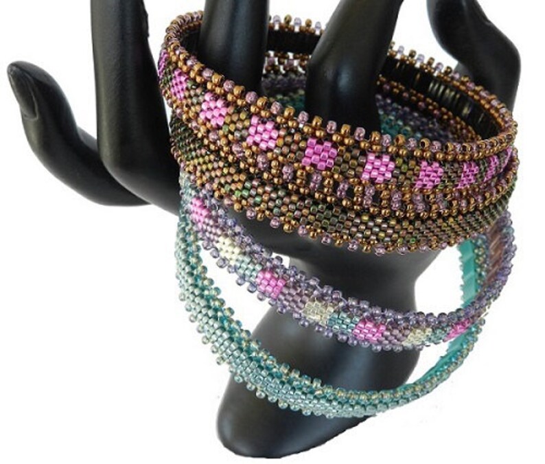 May include: Three beaded bracelets in shades of teal, purple, and brown with pink accents. The bracelets are stacked on a black display stand.
