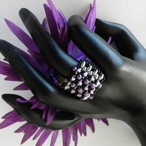 K&ouml;nnte beinhalten: Ein schwarzer Perlenring mit wei&szlig;en und lila Perlen. Der Ring befindet sich an einem schwarzen Handmodell mit einer lila Blume.