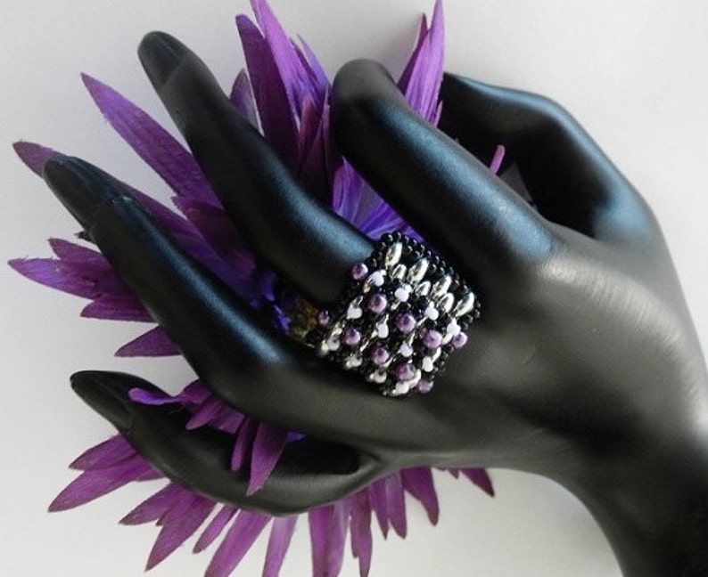 K&ouml;nnte beinhalten: Ein schwarzer Perlenring mit wei&szlig;en und lila Perlen. Der Ring befindet sich an einem schwarzen Handmodell mit einer lila Blume.