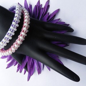 May include: Two beaded bracelets with a gradient of pink, white, and purple beads. The bracelets are on a black mannequin hand with a purple flower behind it.