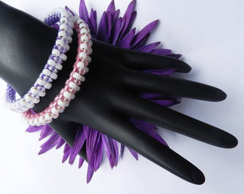 May include: Two beaded bracelets with a gradient of pink, white, and purple beads. The bracelets are on a black mannequin hand with a purple flower behind it.