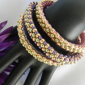 May include: Four beaded bracelets in shades of purple, green, and gold. The bracelets are stacked on top of each other, creating a layered look.
