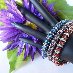 May include: Four beaded bracelets in red, silver, purple, and blue. The bracelets are stacked on a black mannequin hand.