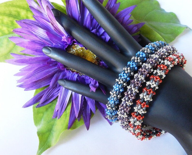 May include: Four beaded bracelets in red, silver, purple, and blue. The bracelets are stacked on a black mannequin hand.