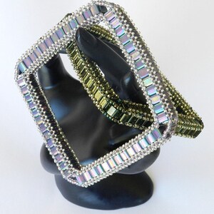 May include: Two beaded bracelets, one silver and one green, are stacked on a black display stand. The bracelets are made of square beads and are iridescent.