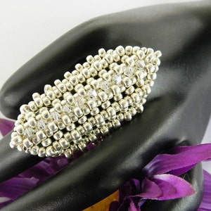 May include: A silver and clear crystal beaded ring. The ring is oval shaped and has a textured design. The ring is on a black hand model.